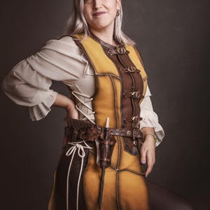 Peut inclure: Une personne porte un gilet en cuir jaune et marron avec une ceinture assortie et une dague. Le gilet a des boucles et des coutures d&eacute;coratives. La personne porte &eacute;galement un chemisier blanc &agrave; manches bouffantes. L'image contient le texte "El Marqu&eacute;s Larp Shop".