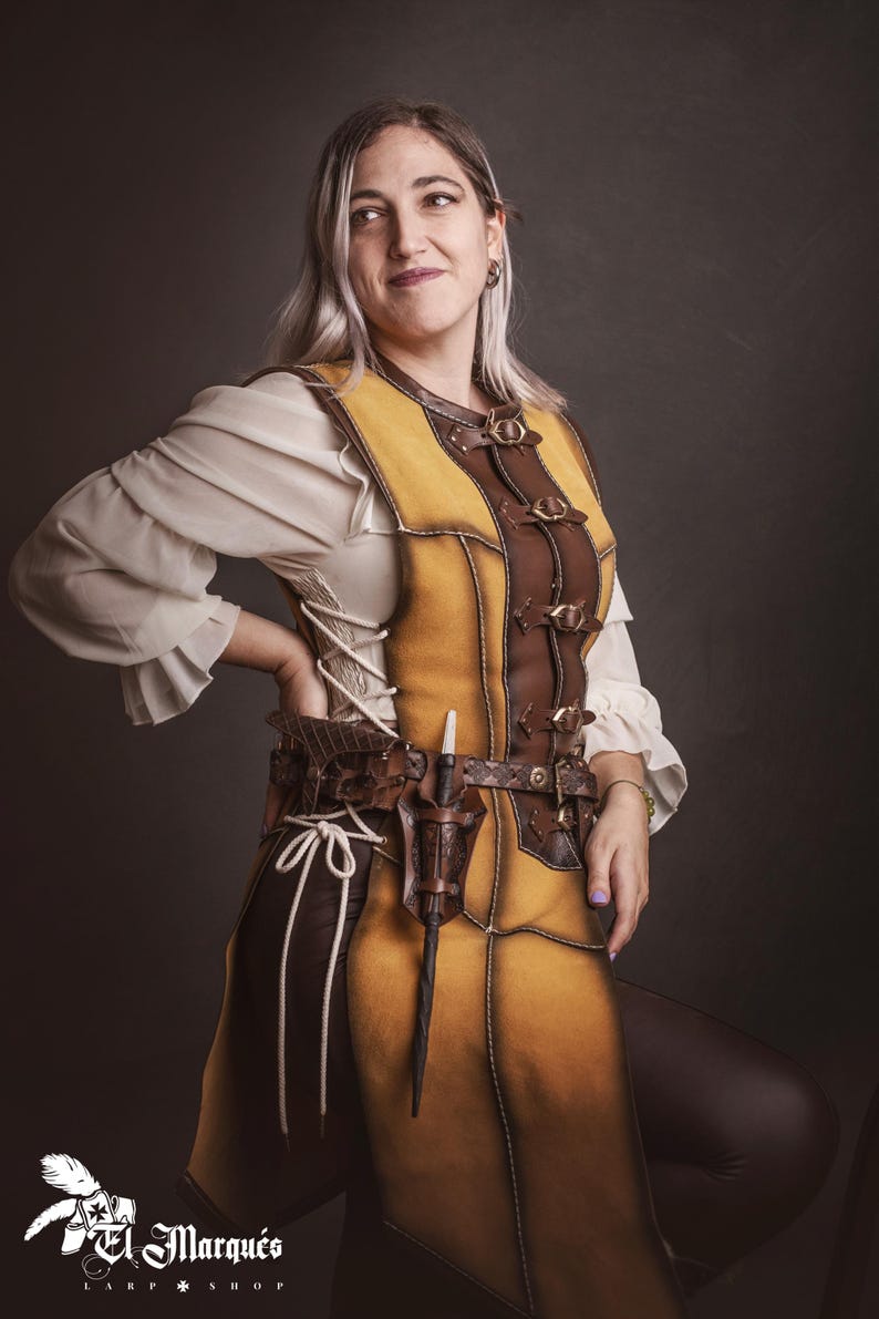 Peut inclure: Une personne porte un gilet en cuir jaune et marron avec une ceinture assortie et une dague. Le gilet a des boucles et des coutures d&eacute;coratives. La personne porte &eacute;galement un chemisier blanc &agrave; manches bouffantes. L'image contient le texte "El Marqu&eacute;s Larp Shop".