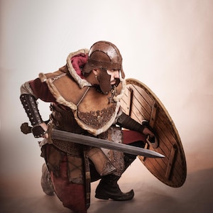 K&ouml;nnte beinhalten: Eine Person in einem braunen Leder- und Fellkost&uuml;m, kniend mit Schwert und Schild. Das Kost&uuml;m umfasst einen Helm, eine R&uuml;stung und Stiefel. Der Schild ist rund und aus Holz. Der Text "El Marqu&eacute;s LARP Shop" ist sichtbar.