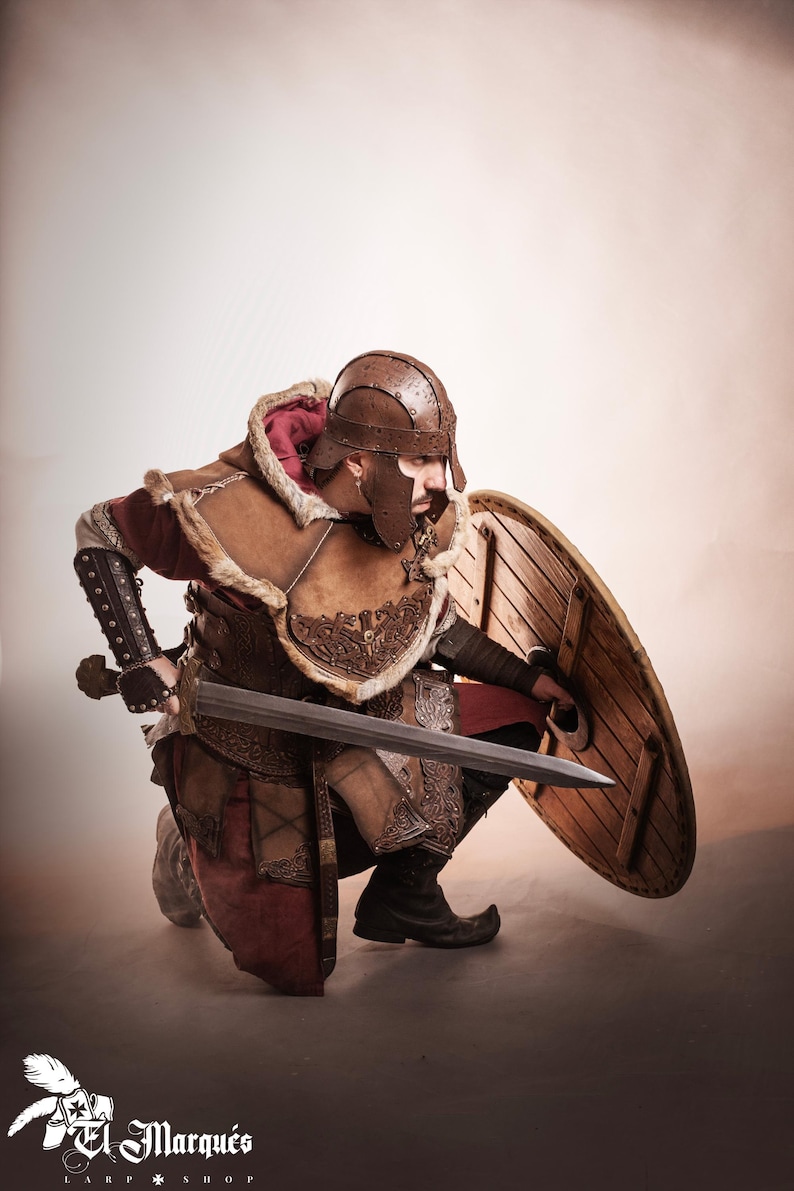 K&ouml;nnte beinhalten: Eine Person in einem braunen Leder- und Fellkost&uuml;m, kniend mit Schwert und Schild. Das Kost&uuml;m umfasst einen Helm, eine R&uuml;stung und Stiefel. Der Schild ist rund und aus Holz. Der Text "El Marqu&eacute;s LARP Shop" ist sichtbar.