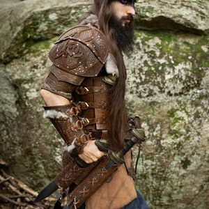 May include: A person wearing brown leather armor with intricate carvings and a fur trim. They are holding two swords with leather sheaths. The person is standing in a forest setting.