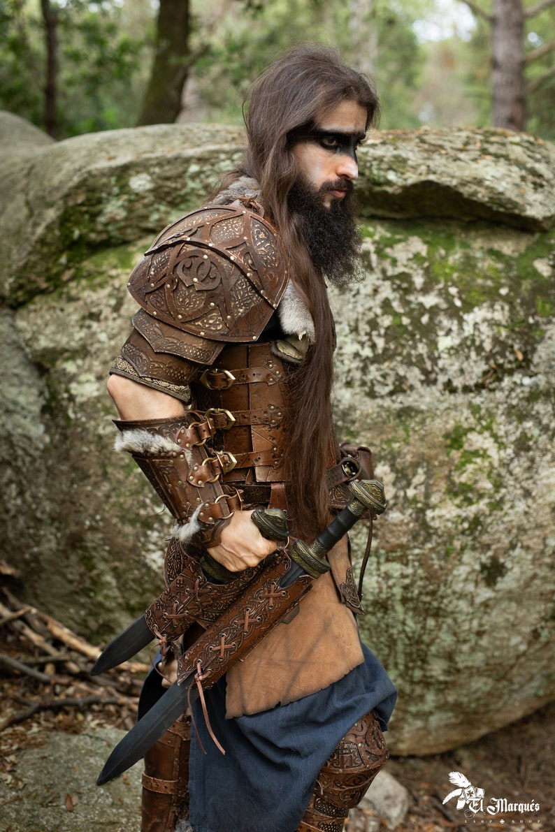 May include: A person wearing brown leather armor with intricate carvings and a fur trim. They are holding two swords with leather sheaths. The person is standing in a forest setting.