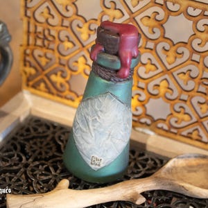 May include: A small, green glass bottle with a white label and a red wax seal. The bottle is sitting on a wooden surface with a decorative pattern.