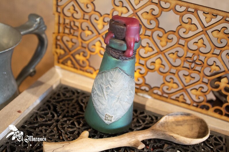 May include: A small, green glass bottle with a white label and a red wax seal. The bottle is sitting on a wooden surface with a decorative pattern.