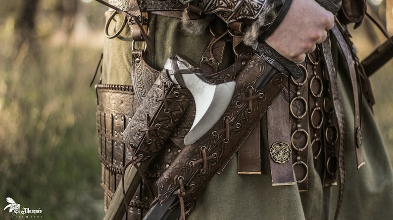 May include: A close-up of a leather belt with intricate carvings and a metal axe attached to it. The belt is worn over a green tunic and has a chainmail belt with metal rings.