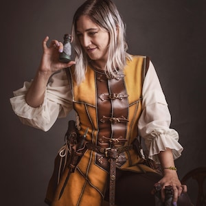 Peut inclure: Une personne porte un gilet en cuir jaune et marron avec une ceinture et un pantalon assortis. Le gilet a des boucles et des coutures d&eacute;coratives. La personne tient une petite bouteille de couleur fonc&eacute;e. L'image est sur fond gris fonc&eacute;.