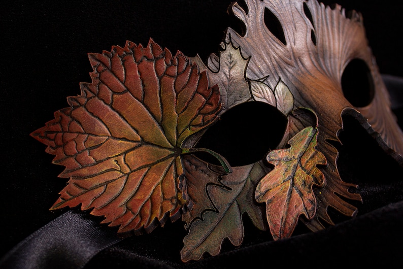 May include: A leather masquerade mask with a design of autumn leaves in shades of brown, green, and gold. The mask has a large leaf on the left side and two smaller leaves on the right side.