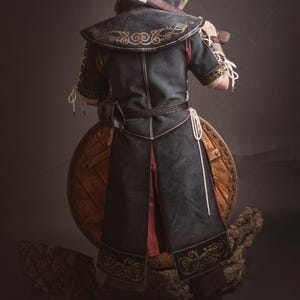 Peut inclure: Costume m&eacute;di&eacute;val avec une cape noire &agrave; capuche, broderies dor&eacute;es et ceinture en cuir. L'ensemble comprend un bouclier en bois et une hache. Id&eacute;al pour le cosplay ou les reconstitutions historiques.