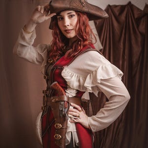Puede incluir: Una mujer con un traje rojo y marr&oacute;n con un sombrero marr&oacute;n y una funda de cuero con una pistola. El traje tiene una blusa blanca con volantes. El fondo es una cortina marr&oacute;n.