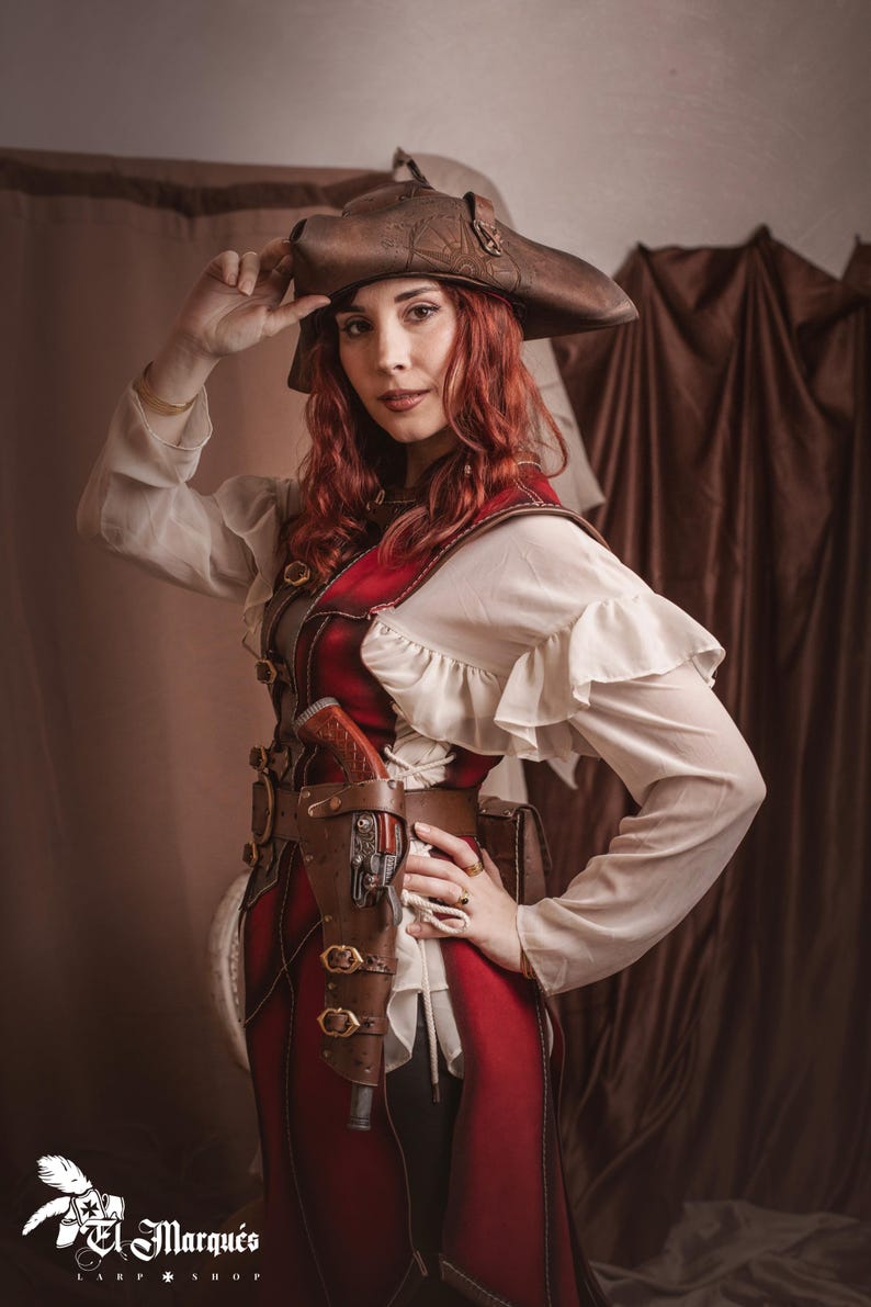 Puede incluir: Una mujer con un traje rojo y marr&oacute;n con un sombrero marr&oacute;n y una funda de cuero con una pistola. El traje tiene una blusa blanca con volantes. El fondo es una cortina marr&oacute;n.