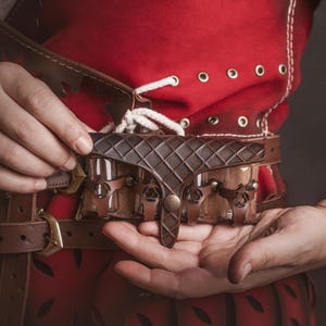 Peut inclure: Une ceinture en cuir marron avec une pochette d&eacute;corative et une boucle. La ceinture est tenue par une personne v&ecirc;tue d'un v&ecirc;tement rouge avec des lacets blancs et des &oelig;illets. La pochette a un motif en losange.