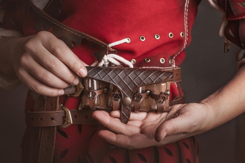 Peut inclure: Une ceinture en cuir marron avec une pochette d&eacute;corative et une boucle. La ceinture est tenue par une personne v&ecirc;tue d'un v&ecirc;tement rouge avec des lacets blancs et des &oelig;illets. La pochette a un motif en losange.
