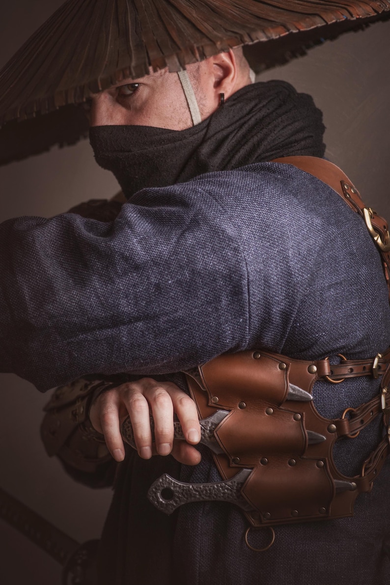 May include: A person wearing a dark blue robe and a wide-brimmed straw hat, holding a silver dagger with a leather sheath. The dagger is attached to a brown leather belt with metal accents.