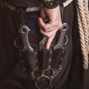 May include: A black leather belt with a metal buckle and two silver throwing knives in a sheath. The belt has a decorative design with metal studs and rings.