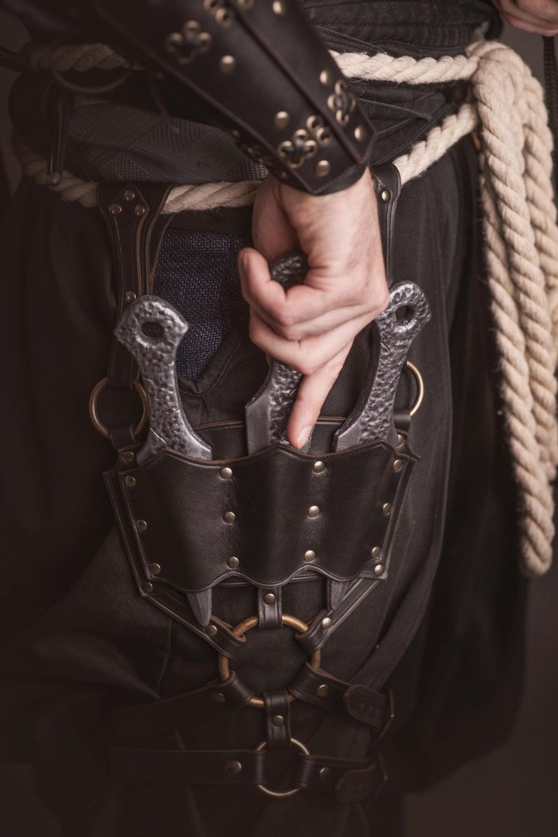 May include: A black leather belt with a metal buckle and two silver throwing knives in a sheath. The belt has a decorative design with metal studs and rings.