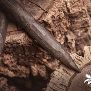 Puede incluir: Primer plano de dos herramientas de madera marrón oscuro, talladas a mano, posiblemente leznas o agujas, sobre una superficie de madera marrón texturizada. Las herramientas tienen un extremo puntiagudo y un diseño tallado y acanalado.