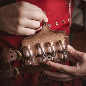 Peut inclure: Une ceinture en cuir marron avec un &eacute;tui assorti contenant quatre petites bouteilles en verre avec des bouchons en li&egrave;ge. La ceinture est port&eacute;e sur un v&ecirc;tement rouge. Les bouteilles sont maintenues par des lani&egrave;res en cuir marron.