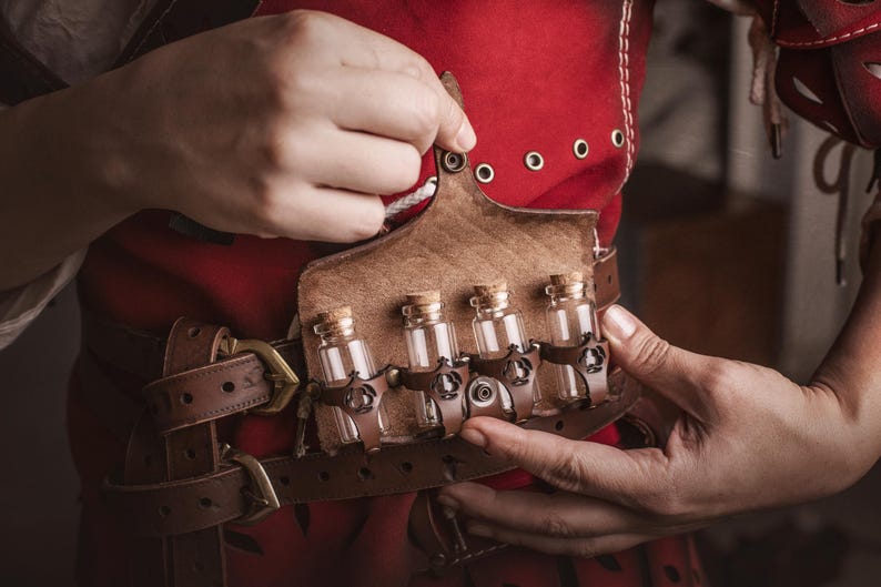 Peut inclure: Une ceinture en cuir marron avec un &eacute;tui assorti contenant quatre petites bouteilles en verre avec des bouchons en li&egrave;ge. La ceinture est port&eacute;e sur un v&ecirc;tement rouge. Les bouteilles sont maintenues par des lani&egrave;res en cuir marron.