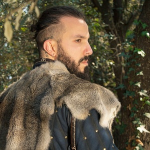 May include: A man with a beard and a shaved head is wearing a fur-lined coat. He is looking to the right and has a serious expression on his face.