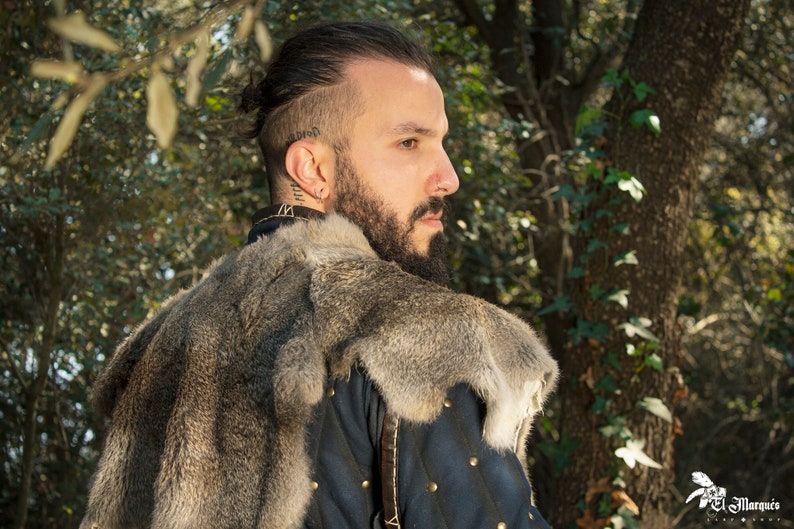 May include: A man with a beard and a shaved head is wearing a fur-lined coat. He is looking to the right and has a serious expression on his face.