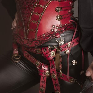 K&ouml;nnte beinhalten: Ein rotes Lederkorsett mit aufwendigen floralen Mustern und goldenen Akzenten. Das Korsett wird &uuml;ber einer schwarzen Hose und einem G&uuml;rtel mit zus&auml;tzlichen Lederriemen und Schnallen getragen.
