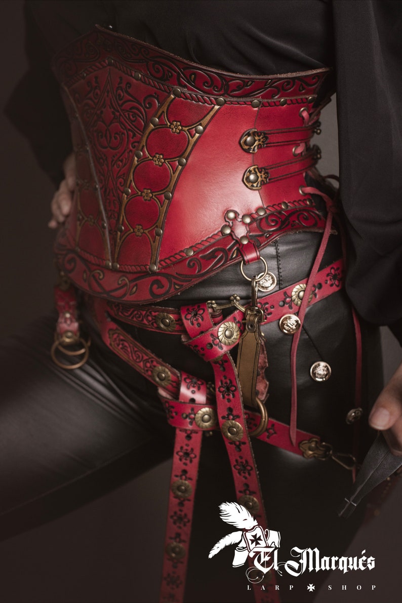 K&ouml;nnte beinhalten: Ein rotes Lederkorsett mit aufwendigen floralen Mustern und goldenen Akzenten. Das Korsett wird &uuml;ber einer schwarzen Hose und einem G&uuml;rtel mit zus&auml;tzlichen Lederriemen und Schnallen getragen.