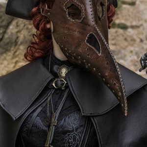May include: A woman wearing a black leather hat, a brown leather plague doctor mask, a black lace top, and a black leather cape. She is also wearing a silver necklace with a locket and a brown leather belt with a silver buckle. The image is taken from the chest up and the background is a blurry stone wall.