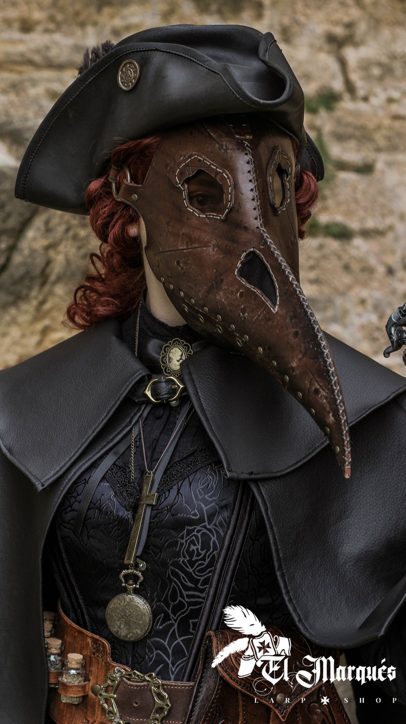 May include: A woman wearing a black leather hat, a brown leather plague doctor mask, a black lace top, and a black leather cape. She is also wearing a silver necklace with a locket and a brown leather belt with a silver buckle. The image is taken from the chest up and the background is a blurry stone wall.