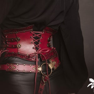 K&ouml;nnte beinhalten: Ein rotes Lederkorsett mit aufwendigen Details und einem Schn&uuml;rr&uuml;cken. Das Korsett hat eine dekorative Metallschnalle und wird &uuml;ber einem schwarzen Hemd getragen.