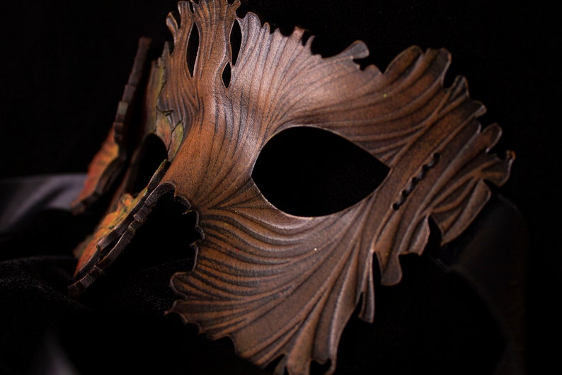 May include: A brown leather masquerade mask with intricate details and a jagged edge. The mask has a dark brown and gold color scheme and is designed to cover one eye.