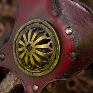 May include: A red leather mask with a gold metal design. The mask has a decorative floral pattern and is secured with metal rivets.