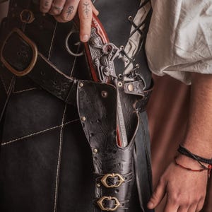 Puede incluir: Una funda de cuero negro con dos hebillas de lat&oacute;n, que contiene una pistola de plata y marr&oacute;n. La funda est&aacute; unida a un cintur&oacute;n de cuero negro con una hebilla de lat&oacute;n.