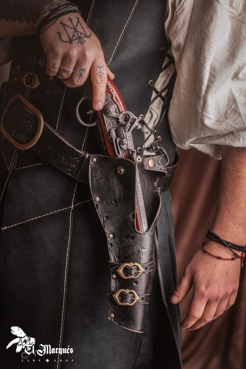 Puede incluir: Una funda de cuero negro con dos hebillas de lat&oacute;n, que contiene una pistola de plata y marr&oacute;n. La funda est&aacute; unida a un cintur&oacute;n de cuero negro con una hebilla de lat&oacute;n.