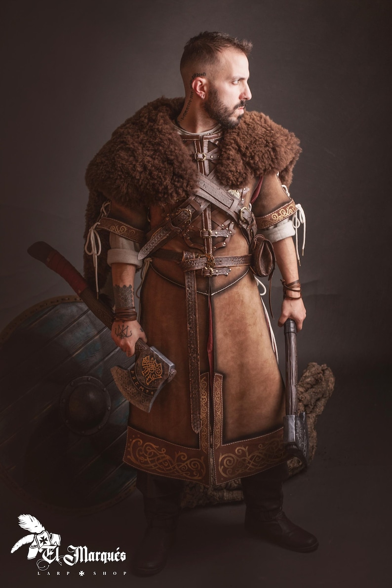 Peut inclure: Costume de Viking en cuir marron avec garniture en fourrure, d&eacute;tails complexes et ceinture d&eacute;corative. L'ensemble comprend une hache, un bouclier et un marteau. Le logo "El Marqu&eacute;s Larp Shop" est visible.