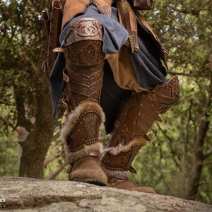 May include: Close-up of a pair of brown leather knee-high boots with intricate metal accents and fur trim. The boots are worn by a person standing on a rock in a forest setting.