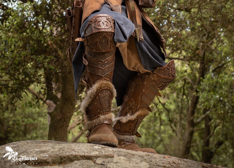 May include: Close-up of a pair of brown leather knee-high boots with intricate metal accents and fur trim. The boots are worn by a person standing on a rock in a forest setting.