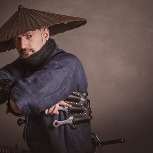 May include: A person wearing a blue shirt and a brown conical hat with a black scarf around their neck. They are holding a silver knife in their right hand and a black leather belt with a silver buckle around their waist.