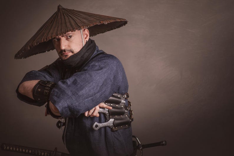 May include: A person wearing a blue shirt and a brown conical hat with a black scarf around their neck. They are holding a silver knife in their right hand and a black leather belt with a silver buckle around their waist.