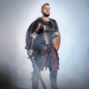Puede incluir: Una persona con armadura medieval negra y roja, sosteniendo una espada y de pie frente a un escudo de madera. La armadura tiene detalles intrincados y está acentuada con oro. La imagen tiene un fondo ahumado.