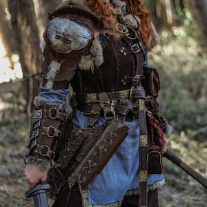 May include: A woman in a forest setting, wearing a brown leather and fur costume with a silver and gold decorative shoulder piece. She is holding a sword in her right hand and has blue face paint.