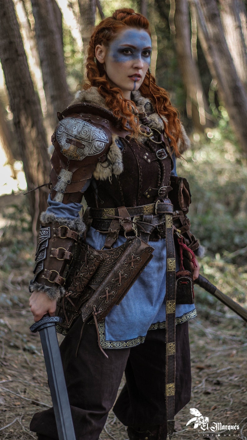 May include: A woman in a forest setting, wearing a brown leather and fur costume with a silver and gold decorative shoulder piece. She is holding a sword in her right hand and has blue face paint.