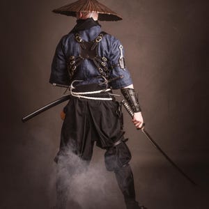 May include: A person wearing a blue and black samurai costume with a straw hat, holding a sword and standing in front of a brown background.