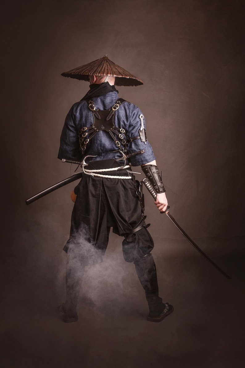 May include: A person wearing a blue and black samurai costume with a straw hat, holding a sword and standing in front of a brown background.