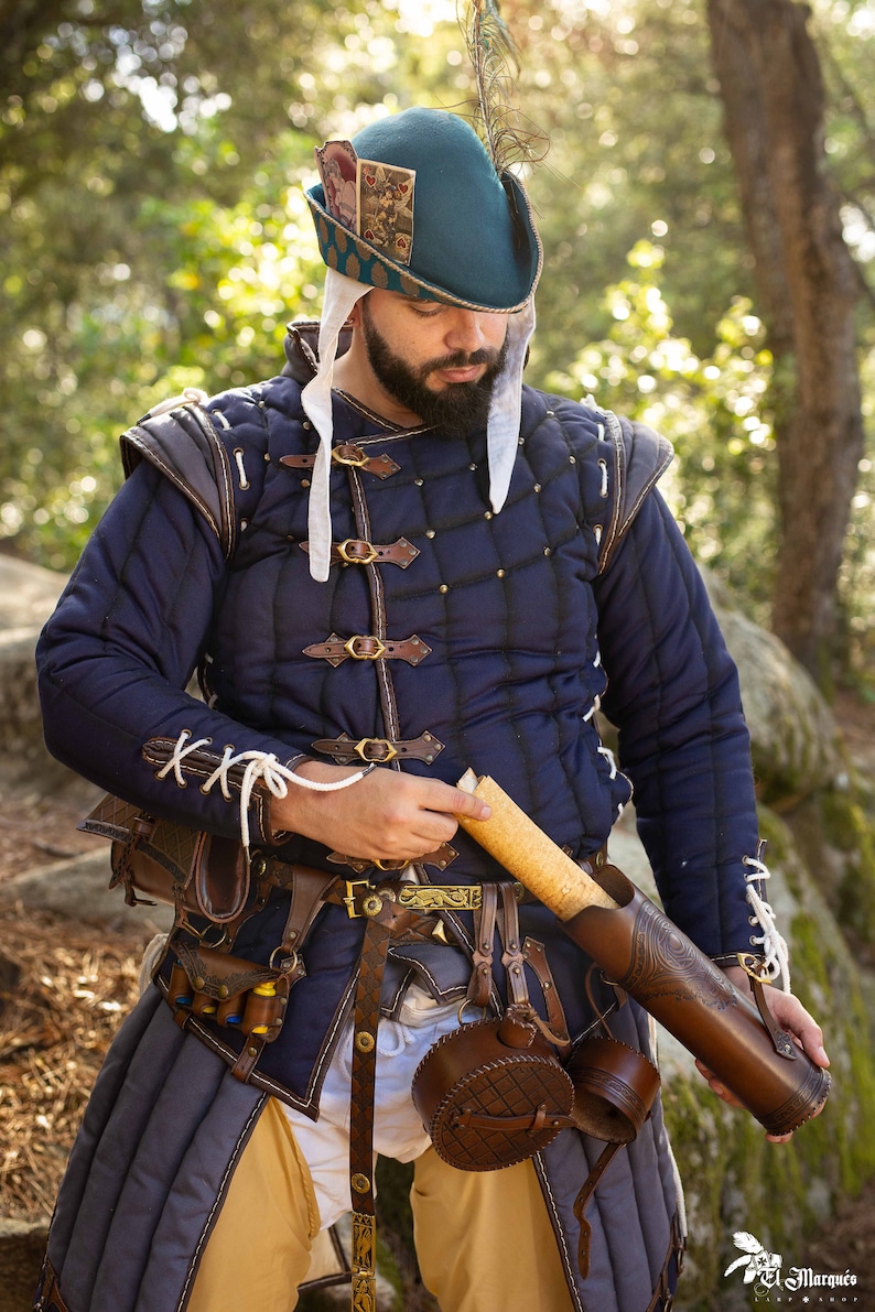 Peut inclure: Un homme portant une veste matelass&eacute;e bleue avec des sangles et des boucles en cuir marron, une chemise en lin blanc et un chapeau vert avec une plume. Il tient un carquois en cuir marron avec une fl&egrave;che en bois &agrave; l'int&eacute;rieur.