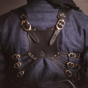 May include: A black leather chest harness with brass buckles and straps. The harness is worn over a dark blue denim shirt.