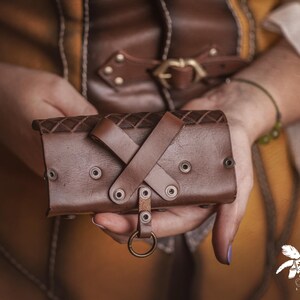 Peut inclure: Un bracelet de poignet en cuir marron avec un dessus &agrave; motif de losanges et des accents rivet&eacute;s. Le bracelet comporte une conception &agrave; lani&egrave;res crois&eacute;es et un anneau en m&eacute;tal. L'image montre le bracelet tenu.