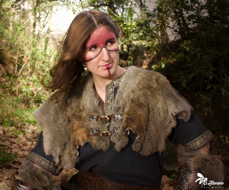 May include: A woman with brown hair and red and black face paint wears a dark blue tunic with a fur collar and matching fur arm warmers. The tunic has a gold trim and buckles.
