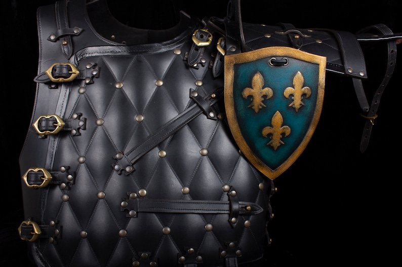 K&ouml;nnte beinhalten: Schwarze Leder-R&uuml;stung mit gestepptem Design und goldenen Schnallen. Ein gold-blauer Schild mit drei goldenen Lilien ist an der R&uuml;stung befestigt.