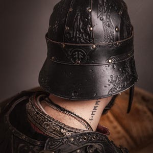 K&ouml;nnte beinhalten: Schwarzer Lederhelm mit gepr&auml;gten Designs und Messingnieten. Der Helm hat einen Nackenschutz und ein dekoratives Band. Das Bild zeigt die R&uuml;ckseite des Helms und einen Teil der R&uuml;stung des Tr&auml;gers. Der Helm ist von einem LARP-Shop.