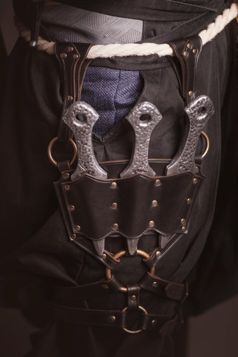May include: A leather belt with a metal buckle and three silver throwing knives attached to it.
